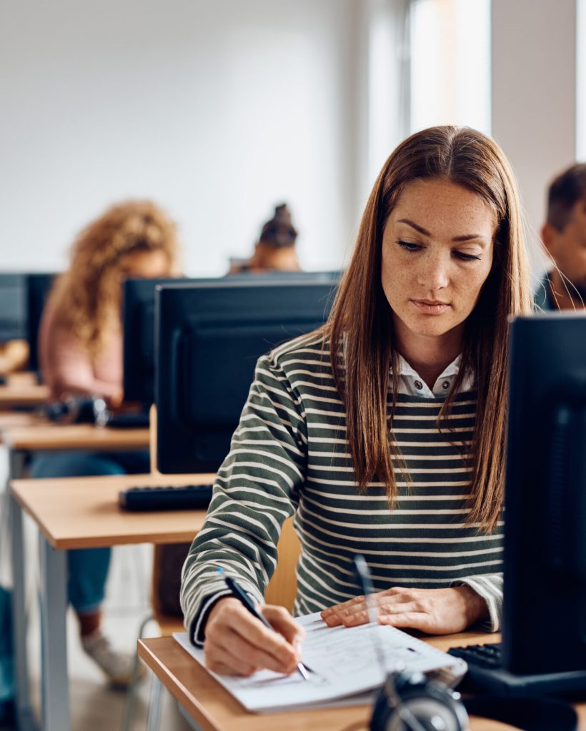 Mid adult woman using desktop PC and taking notes during computer class in the classroom.